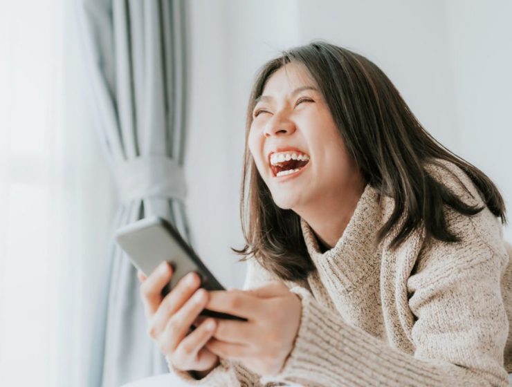 Happy Asian woman laughing while lying on the bed using smartphone mobile at home